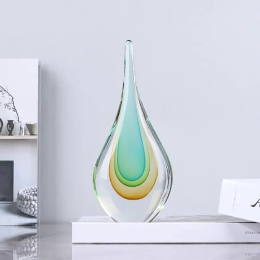 Hand-blown clear glass teardrop sculpture with aqua and amber layers on a desk