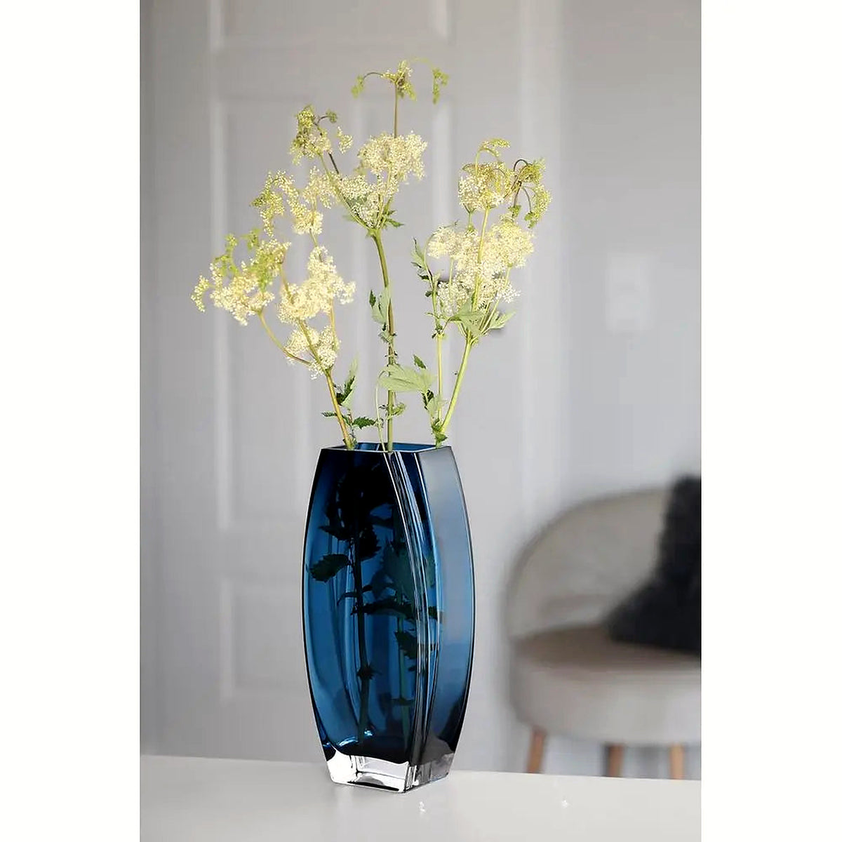 Heavy blue-teal art glass vase with tall green filler stems on a white table (stems and décor shown for styling)