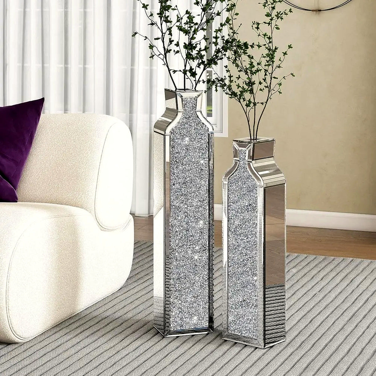 Two tall mirrored floor vases with crushed-crystal inlay holding slender branches beside a cream armchair