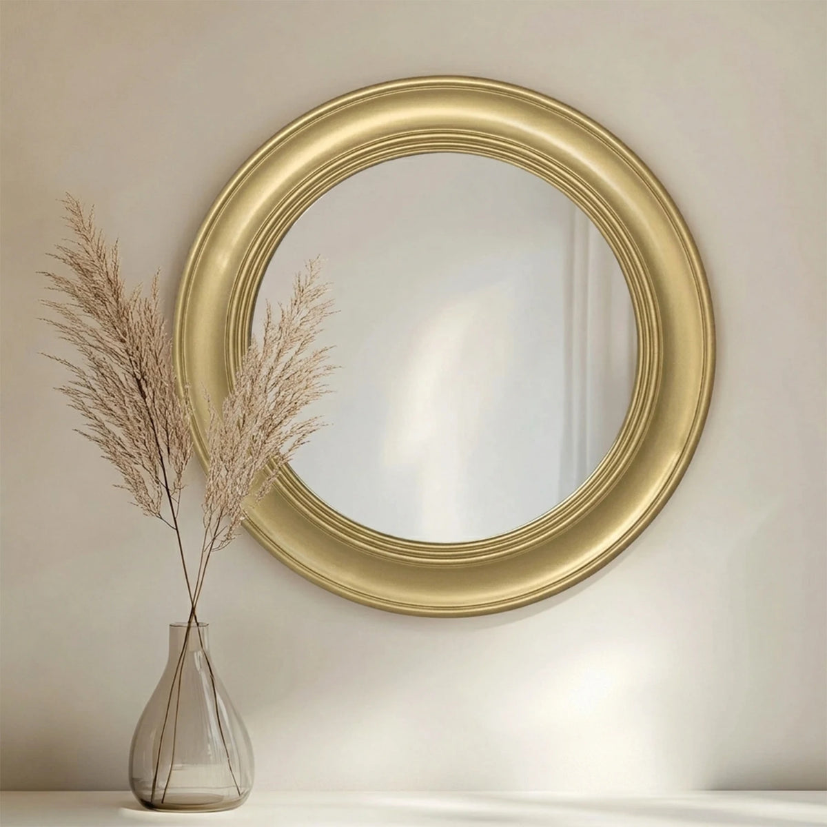 Brushed gold round mirror on a wall beside a glass vase of dried grass