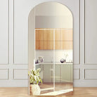 Large arched floor mirror with a slim pale natural wood frame leaning against a white paneled wall