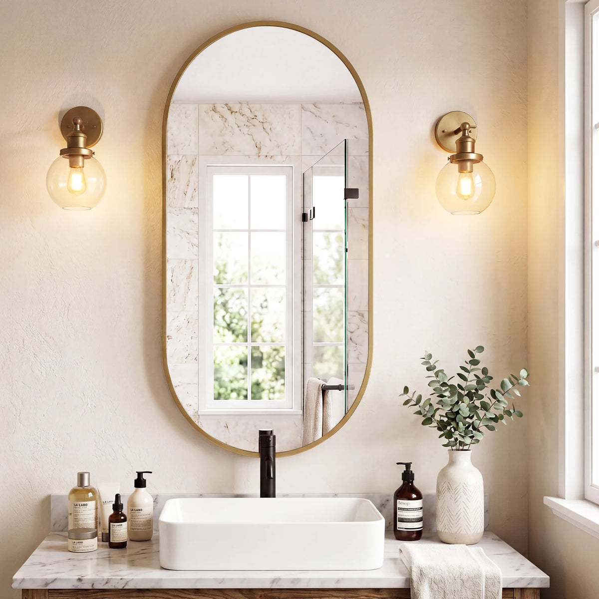 Oval pill-shaped metal frame bathroom mirror above a vessel sink with globe sconces