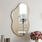 Gold wavy asymmetrical wall mirror hung vertically above pink flowers