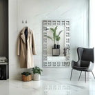 Greek-key Venetian wall mirror hung vertically in a white entryway beside a coat hook
