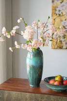 Teal ceramic vase with pink blossoms beside a fruit bowl