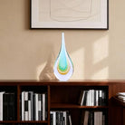 Hand-blown clear glass teardrop sculpture with aqua and amber layers on a bookshelf