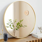 Round matte gold metal frame mirror leaning on a wood shelf beside a green branch