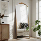 Ornate carved floor mirror with slim gold frame leaning beside a rattan cabinet