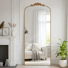 Arched gold standing floor mirror with carved crest leaning in a white living room