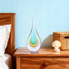 Hand-blown clear glass teardrop sculpture with aqua and amber layers on a bedside table