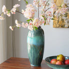 Teal reactive-glaze ceramic vase with blossom stems on a wood sideboard