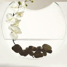 Close-up of the clear ellipse vessel holding orchid stems and dark river stones