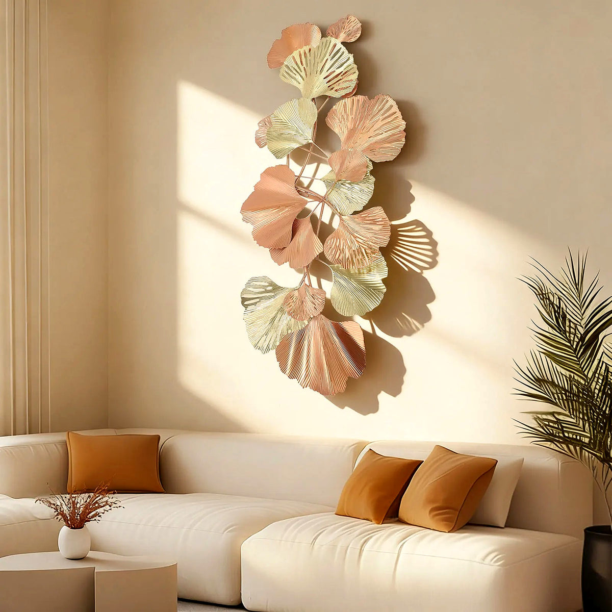 Large ginkgo leaf metal wall art in blush, gold and sage, hung vertically above a cream sofa in warm sunlight
