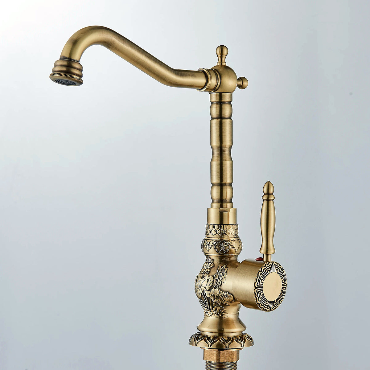 Carved antique brass swivel basin faucet with floral relief, shown full-length against a soft grey studio backdrop