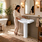 Glossy white pedestal sink in a sunlit bath with a freestanding tub, brass accents and soft patterned flooring