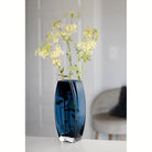 Heavy blue-teal art glass vase with tall green filler stems on a white table (stems and décor shown for styling)