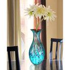 Hand-blown teal art glass vase on a dining table with white star-shaped blooms (flowers and furniture shown for styling)
